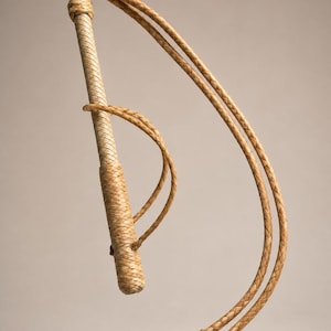 May include: A tan leather whip featuring a braided handle and a long, flexible braided lash that curves outward. The handle includes a small braided loop attached near the top, and the end of the lash terminates in several thin, loose leather strips.