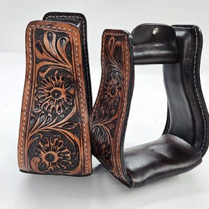 May include: A pair of brown leather stirrups with intricate floral tooling. The stirrups feature a dark brown interior and edges, with lighter brown and black floral designs. White stitching accents the edges.