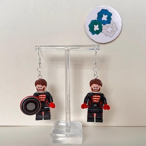Op de afbeelding: Een paar handgemaakte oorbellen met miniatuur Lego-figuren. Elke figuur draagt een zwart en rood uniform, met een bruine baard en haar. Eén figuur houdt een zwart en rood schild vast. De oorbellen hangen aan zilverkleurige haken.