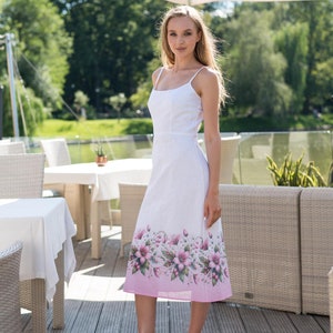 May include: White midi dress with spaghetti straps and a floral print border. The dress features a fitted bodice and a flared skirt. The floral design includes pink and purple flowers on a light pink background. The dress is approximately 40 inches long.