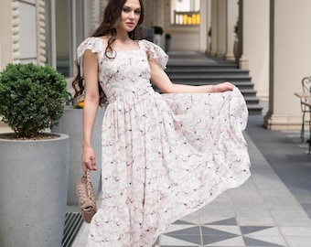 May include: A white midi dress with a pink floral pattern. The dress features a square neckline, flutter sleeves, and a tiered skirt. The woman is wearing nude heels and carrying a small, woven handbag.