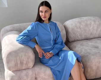 Könnte beinhalten: Eine Frau in einem hellblauen Kleid mit weißem Spitzenbesatz sitzt auf einem hellrosa Sofa. Das Kleid hat lange Ärmel und einen runden Ausschnitt.