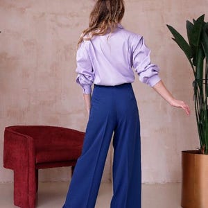 May include: A person wearing a light purple button-down shirt with long sleeves and blue wide-leg pants. The outfit is paired with black heels. A red chair and a potted plant are in the background.