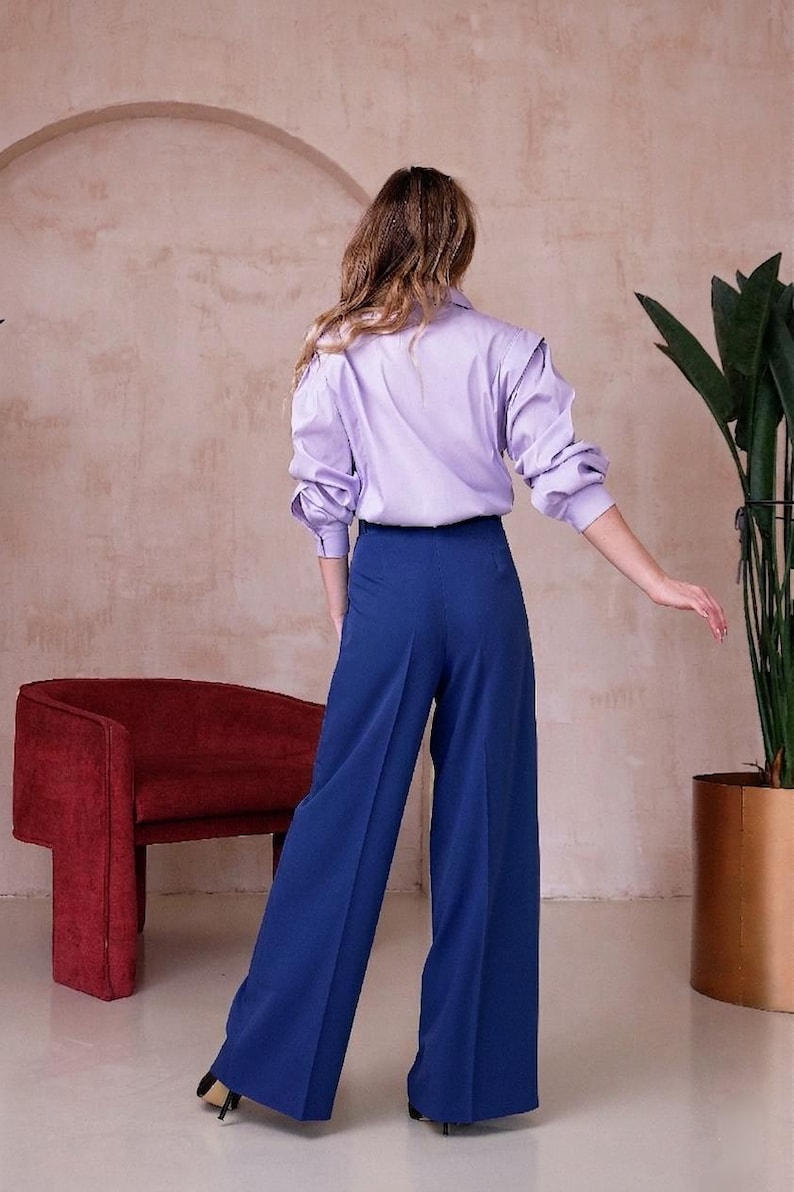 May include: A person wearing a light purple button-down shirt with long sleeves and blue wide-leg pants. The outfit is paired with black heels. A red chair and a potted plant are in the background.