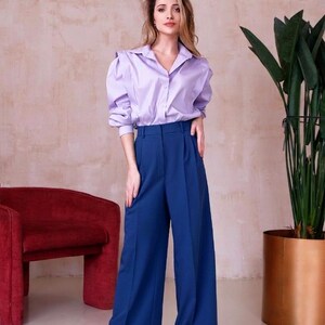 May include: A woman wearing a light purple button-down shirt with puffed sleeves and high-waisted, wide-leg blue pants. The outfit is paired with black shoes. A red armchair and a potted plant are in the background.