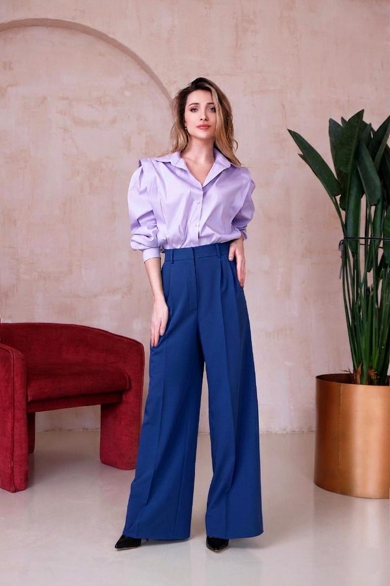 May include: A woman wearing a light purple button-down shirt with puffed sleeves and high-waisted, wide-leg blue pants. The outfit is paired with black shoes. A red armchair and a potted plant are in the background.