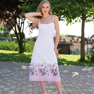 May include: A white midi dress with thin shoulder straps and a square neckline. The lower portion of the skirt features a printed floral pattern in shades of pink and green, transitioning into a solid pink gradient at the hemline.
