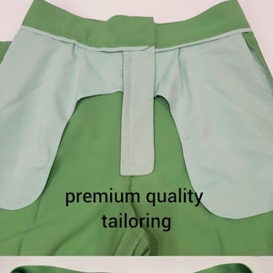 May include: Close-up of green pants with details of hand-stitched hem and premium quality tailoring. The pants have a light green interior lining. The brand tag is visible. The pants are a solid color.