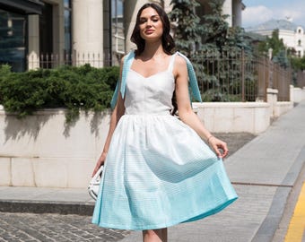 May include: A woman wearing a white and light blue ombre dress with thin straps that tie at the shoulders. She is walking on a sidewalk in front of a building.