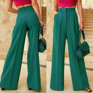 May include: Emerald green wide-leg trousers with a high waist and pleats. The trousers are paired with a matching green handbag. The model is wearing a pink crop top and black sandals. The trousers have a classic, tailored design.