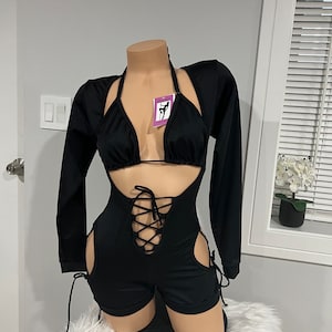 Pode incluir: Macacão preto com cordões na frente e recortes nas laterais. O macacão tem mangas compridas e decote halter. É combinado com um top curto preto com decote halter.