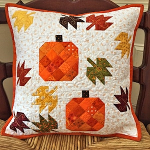 May include: A white pillow with an orange and brown patchwork pumpkin design and green and brown leaf designs. The pillow has a white background with a small floral pattern and an orange border.