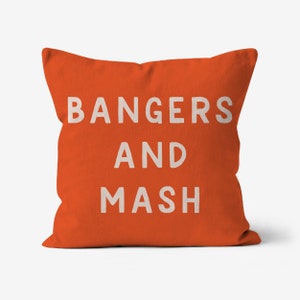 May include: Square orange pillow with the words "BANGERS AND MASH" printed in white block letters. The pillow is a solid orange color and has a soft, textured appearance. The text is centered on the pillow.