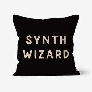 Może przedstawiać: Czarna kwadratowa poduszka z napisem "SYNTH WIZARD" w jasnobeżowym kolorze. Ta dekoracyjna poduszka jest przeznaczona na sofę lub łóżko i ma minimalistyczny, nowoczesny design.