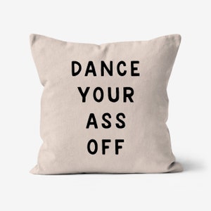 Może przedstawiać: Kwadratowa, beżowa poduszka z napisem "DANCE YOUR ASS OFF" wydrukowanym pogrubioną, czarną czcionką. Poduszka ma prosty, nowoczesny design i neutralną kolorystykę.