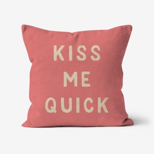 May include: A square cushion in a coral hue, featuring the text "KISS ME QUICK" in a light beige font. This decorative cushion is designed for a bed or sofa.