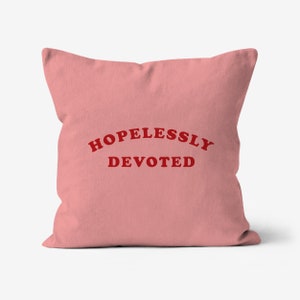 Könnte beinhalten: Ein quadratisches, hellrosa Kissen mit dem roten Schriftzug "HOPELESSLY DEVOTED" in einem Bogen. Das Kissen hat einen einfarbigen Hintergrund und eine einfache, auffällige Botschaft.