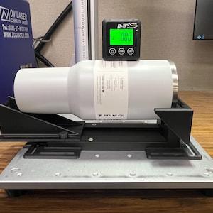 May include: A white Stanley tumbler is secured in a black and silver measuring device. A digital gauge displays "000" in green. The setup is on a wooden surface, with a blue OV Laser machine visible in the background.