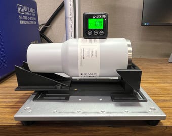 May include: A white Stanley tumbler is secured in a black and silver measuring device. A digital gauge displays "000" in green. The setup is on a wooden surface, with a blue OV Laser machine visible in the background.