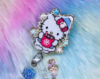 May include: A sparkly blue glitter badge reel with a Hello Kitty character holding a red mug with a snowflake design. The character is wearing a blue and white Santa hat. The background is a fuzzy pink and blue surface.