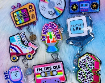 Op de afbeelding: Negen kleurrijke, glinsterende, 90's-thema badge-rollen met verschillende ontwerpen, waaronder een retro-tv, rol schaatsen, een lava lamp, een cassettebandje, een mobiele telefoon, een ghettoblaster, een computer en een letter 'S' met een luipaardprint.
