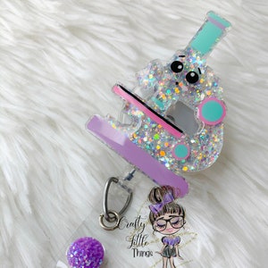 May include: A clear purple and pink glitter microscope badge reel with a cute cartoon face. The reel has a silver swivel clip and a purple glitter charm. The text "Crafty Little Things" is visible on the badge reel.