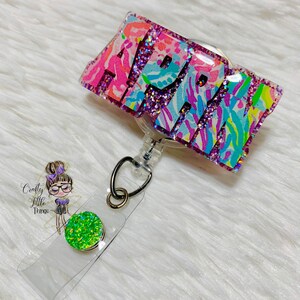 May include: A pink, blue, and green glitter badge holder with the name "APRIL" in white letters. The badge holder is attached to a clear retractable badge reel with a green glitter button.