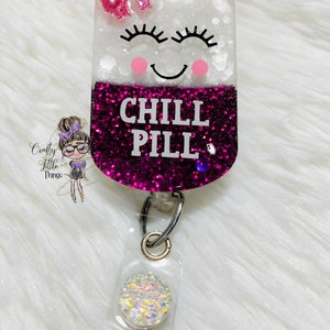 May include: A pink and white glitter pill shaped badge holder with a smiling face, eyelashes, and the text "CHILL PILL". The badge holder has a clear plastic retractable badge reel with a sparkly round charm.