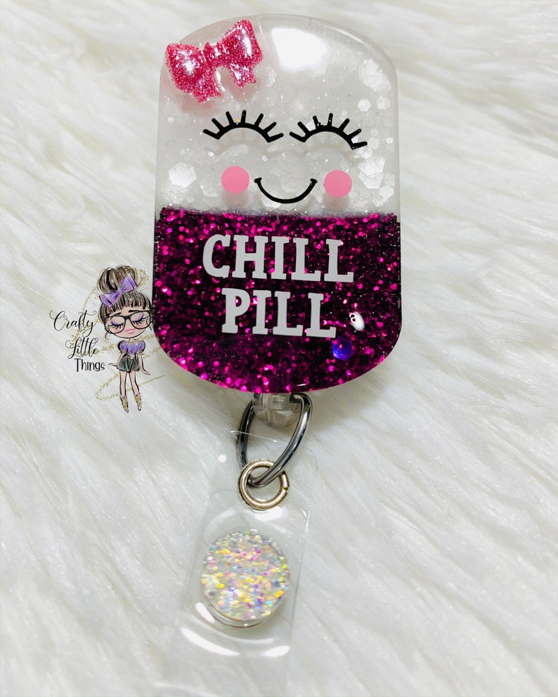 May include: A pink and white glitter pill shaped badge holder with a smiling face, eyelashes, and the text "CHILL PILL". The badge holder has a clear plastic retractable badge reel with a sparkly round charm.