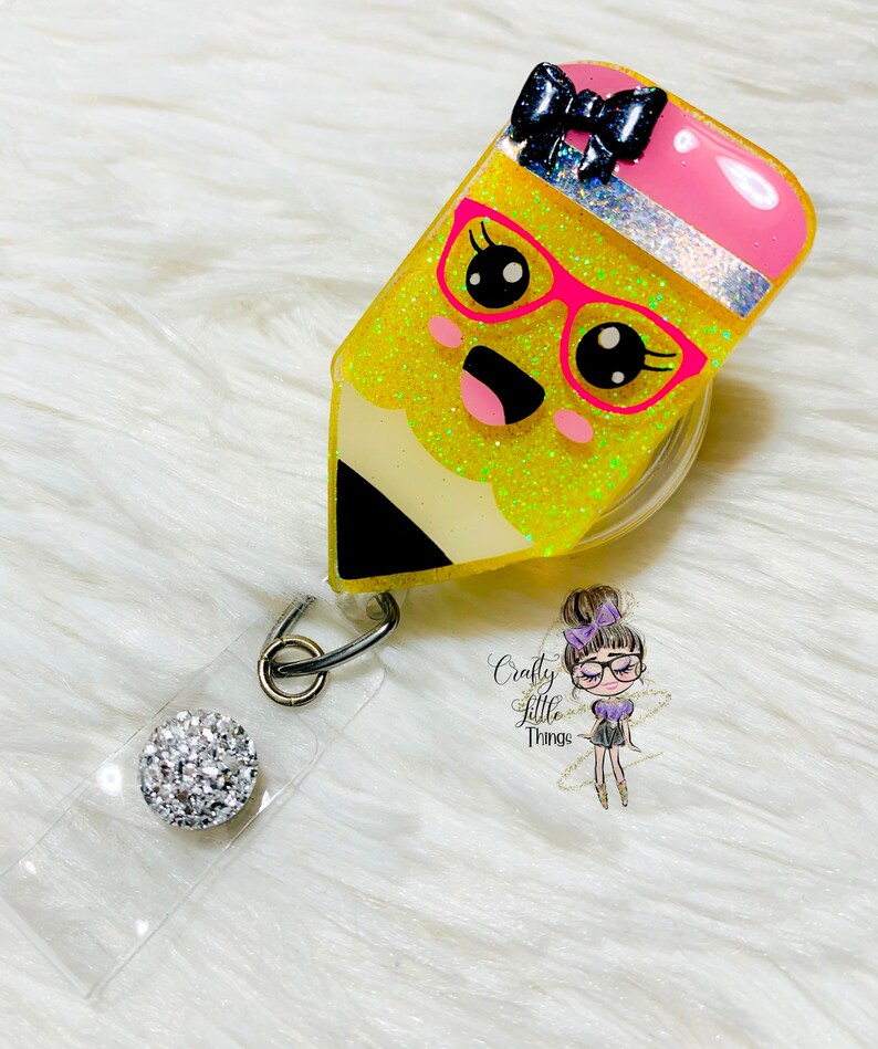 May include: Yellow glitter pencil charm with a pink eraser, black bow, and pink glasses. The charm is attached to a clear badge holder with a silver rhinestone button.