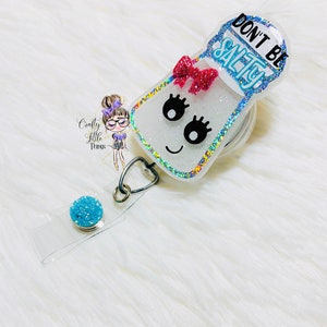 May include: A white glitter salt shaker with a blue rim and a pink bow. The shaker has a black face with eyelashes and a smiling mouth. The text "Don't Be Salty" is written in blue on the top of the shaker. The shaker is attached to a clear retractable badge reel with a blue glitter charm.