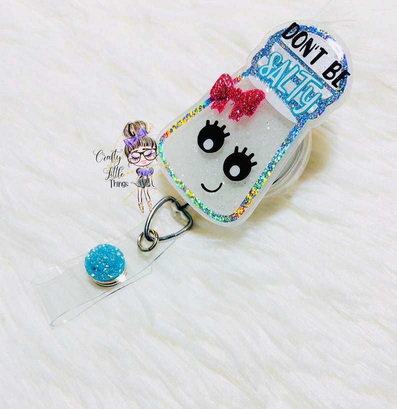 May include: A white glitter salt shaker with a blue rim and a pink bow. The shaker has a black face with eyelashes and a smiling mouth. The text "Don't Be Salty" is written in blue on the top of the shaker. The shaker is attached to a clear retractable badge reel with a blue glitter charm.