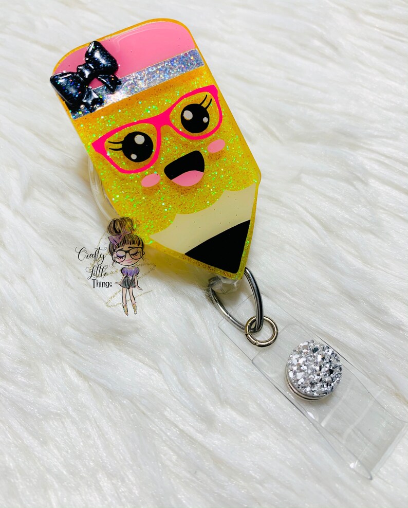 May include: A yellow glitter pencil with a pink eraser, black bow, and pink glasses. The pencil has a happy face and is attached to a clear badge holder with a silver clip and a clear rhinestone.
