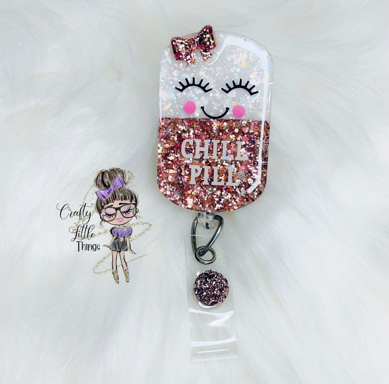 May include: A pink and white glitter resin badge holder with a smiling face and the text "CHILL PILL". The holder is attached to a clear plastic retractable badge reel with a pink glitter button.