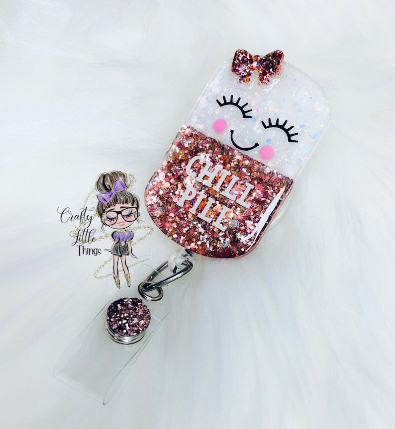 May include: A pink and white glitter resin badge holder with a smiling face and the words "Chill Pill" in white lettering. The badge holder is attached to a clear plastic badge holder with a silver clip.