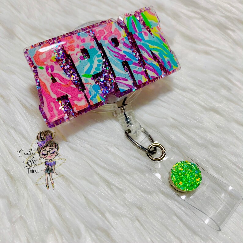 May include: A pink, blue, and green glitter badge holder with the name "APRIL" in large letters. The badge holder is attached to a clear retractable badge reel with a green glitter button.