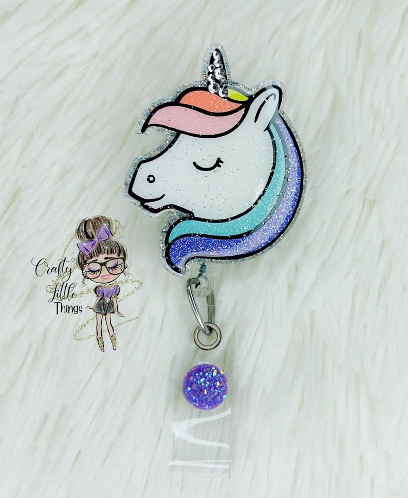 May include: A sparkly white unicorn head with a rainbow mane and a silver glitter background. The unicorn is attached to a retractable badge holder with a purple glitter button and a clear plastic badge holder.