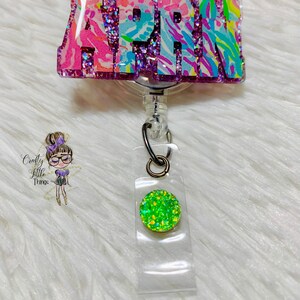 May include: A pink, blue, and green glitter badge holder with the name "APRIL" on it. The badge holder has a retractable cord and a green glitter charm.
