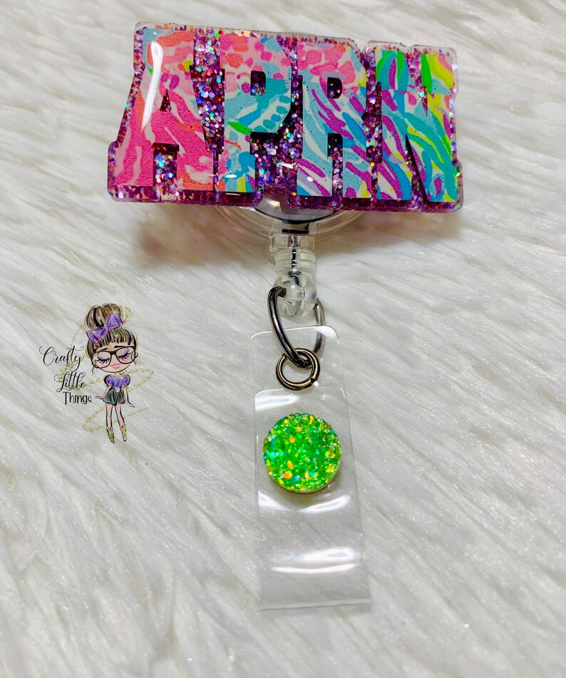 May include: A pink, blue, and green glitter badge holder with the name "APRIL" on it. The badge holder has a retractable cord and a green glitter charm.