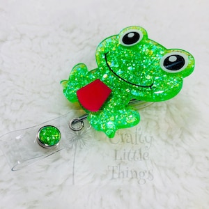 May include: A green glitter frog badge reel with a red accent. The frog has large black and white eyes and a smiling mouth. The badge reel has a clear plastic holder and a green glitter button.