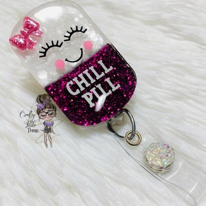 May include: A clear resin badge holder with a pink glitter filled pill shape. The pill has a white face with a smiling cartoon face and the words "CHILL PILL" in black text.