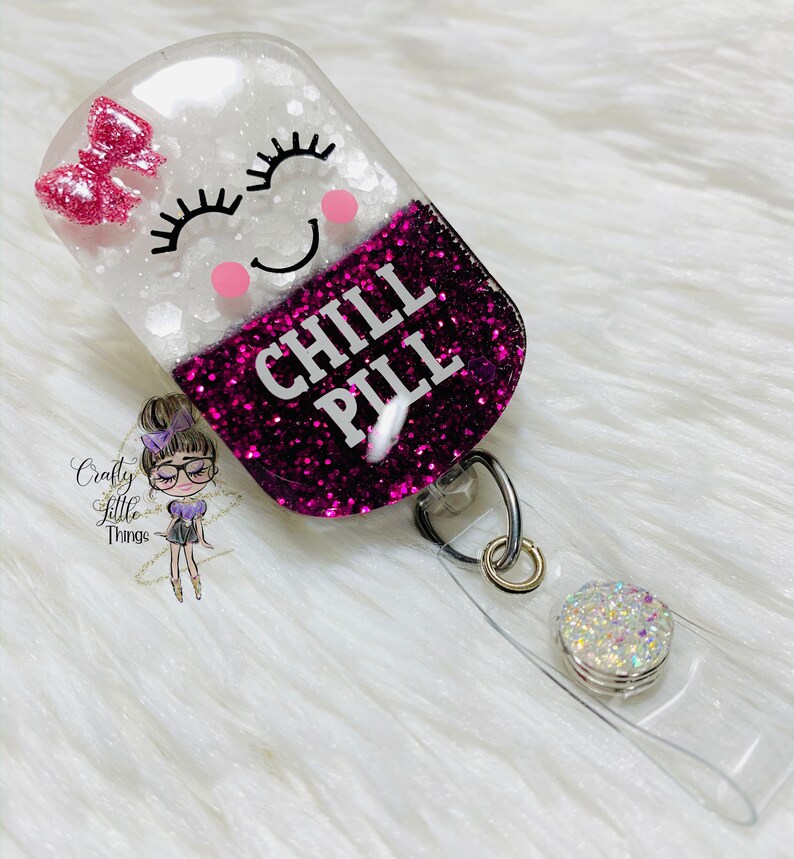 May include: A clear resin badge holder with a pink glitter filled pill shape. The pill has a white face with a smiling cartoon face and the words "CHILL PILL" in black text.