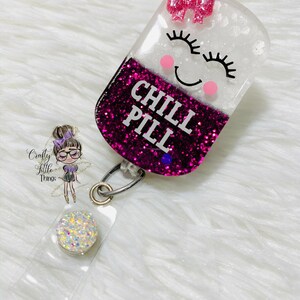 May include: A pink and white glitter resin badge holder shaped like a pill with a smiling face and the text "CHILL PILL". The badge holder has a clear plastic strap and a silver swivel clip.
