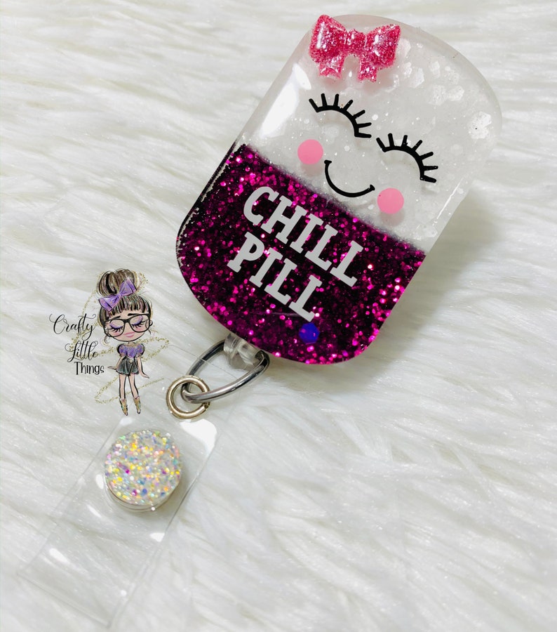 May include: A pink and white glitter resin badge holder shaped like a pill with a smiling face and the text "CHILL PILL". The badge holder has a clear plastic strap and a silver swivel clip.