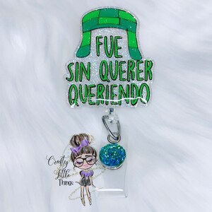 May include: A green and white glitter badge holder with a cartoon girl wearing glasses and a green and blue glitter charm. The badge holder has the text "FUE SIN QUERER QUERIENDO" on it.