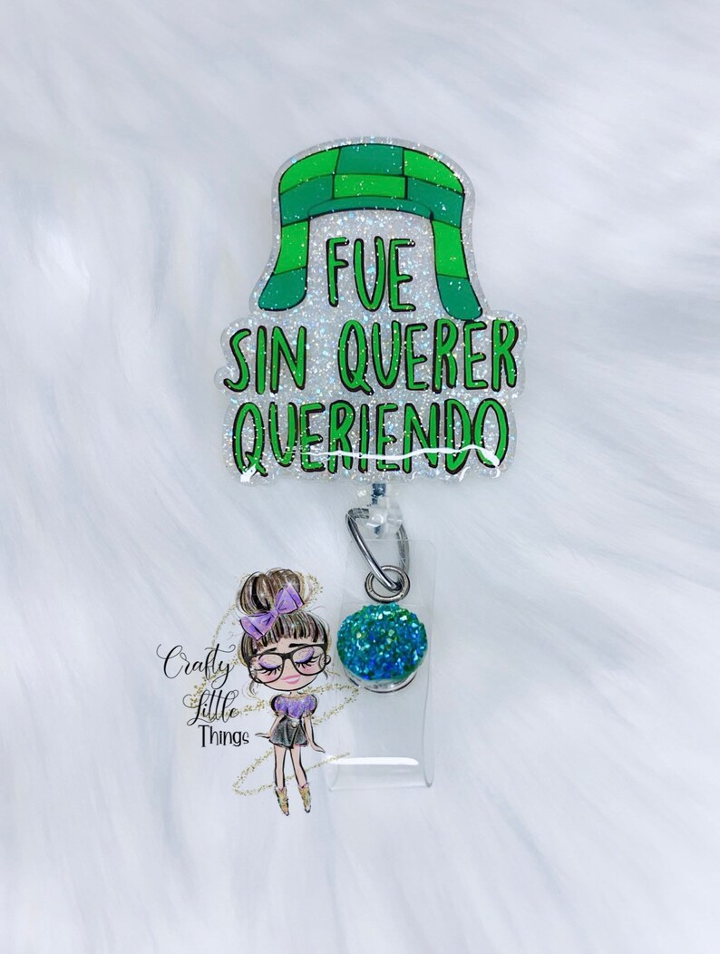 May include: A green and white glitter badge holder with a cartoon girl wearing glasses and a green and blue glitter charm. The badge holder has the text "FUE SIN QUERER QUERIENDO" on it.