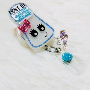 May include: A white salt shaker with a pink bow and glitter, with the text "Don't Be Salty" on the front. The shaker is attached to a clear retractable badge holder with a blue glitter charm.