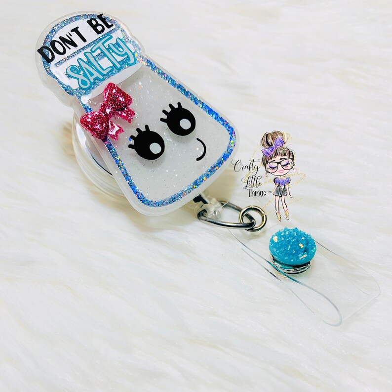 May include: A white salt shaker with a pink bow and glitter, with the text "Don't Be Salty" on the front. The shaker is attached to a clear retractable badge holder with a blue glitter charm.