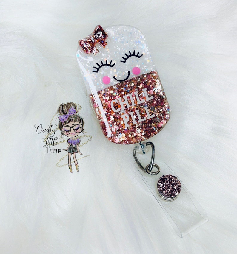 May include: A pink and white glitter resin badge holder with a smiling pill design. The pill has a pink bow and the text "CHILL PILL" in black lettering.