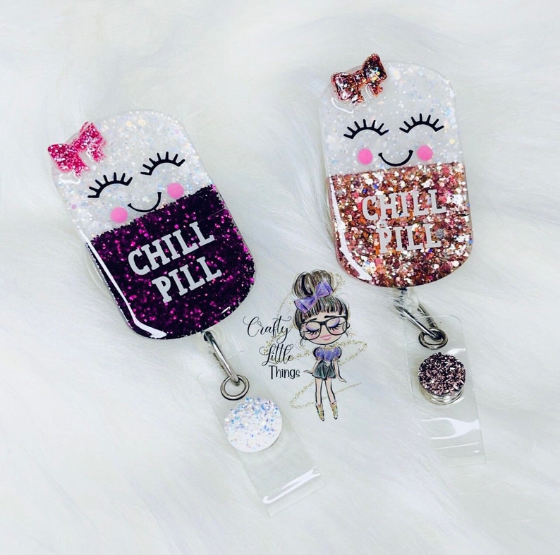 May include: Two glitter filled pill shaped resin keychains with a smiling face and the text "Chill Pill". The keychains are attached to clear retractable badge holders with glitter accents.